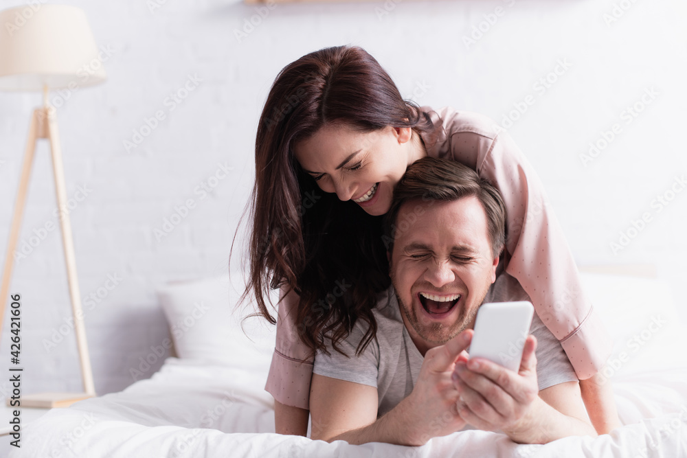 Laughing man holding cellphone on blurred foreground near wife on bed in morning Laughing man holding cellphone on blurred foreground near wife on bed in morning
