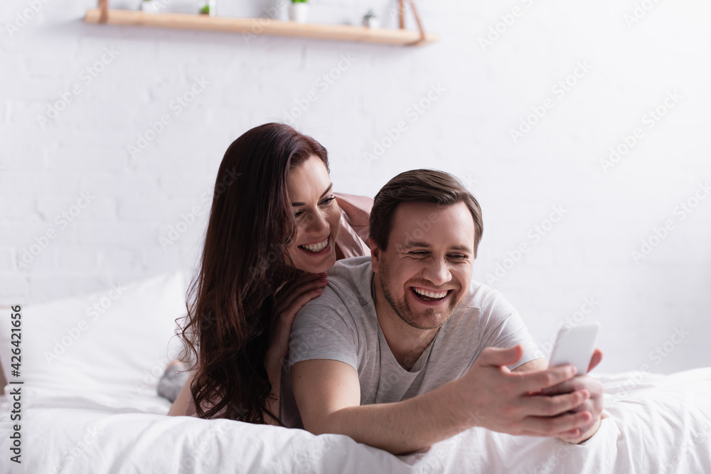 Smiling man using smartphone on blurred foreground near cheerful wife on bed Smiling man using smartphone on blurred foreground near cheerful wife on bed