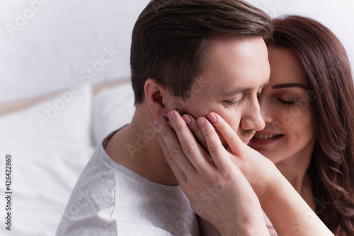 Smiling woman on blurred background touching cheek of husband in bedroom