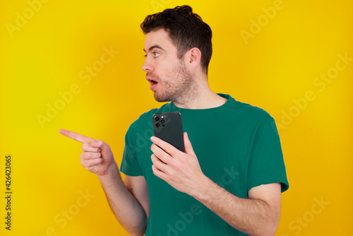 Stunned young handsome caucasian man wearing green t-shirt against yellow background points sideways right copy space, recommends product, sees astonishing thing