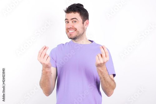 young handsome caucasian man wearing purple t-shirt against white background keeps palms together, has pleased expression. Glad attractive male makes request, pleads for mercy. Hopeful young adult.