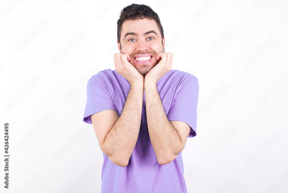 Dreamy young handsome caucasian man wearing purple t-shirt against white background keeps hands pressed together under chin, looks with happy expression, has toothy smile. Dreamy young handsome caucasian man wearing purple t-shirt against white background keeps hands pressed together under chin, looks with happy expression, has toothy smile.