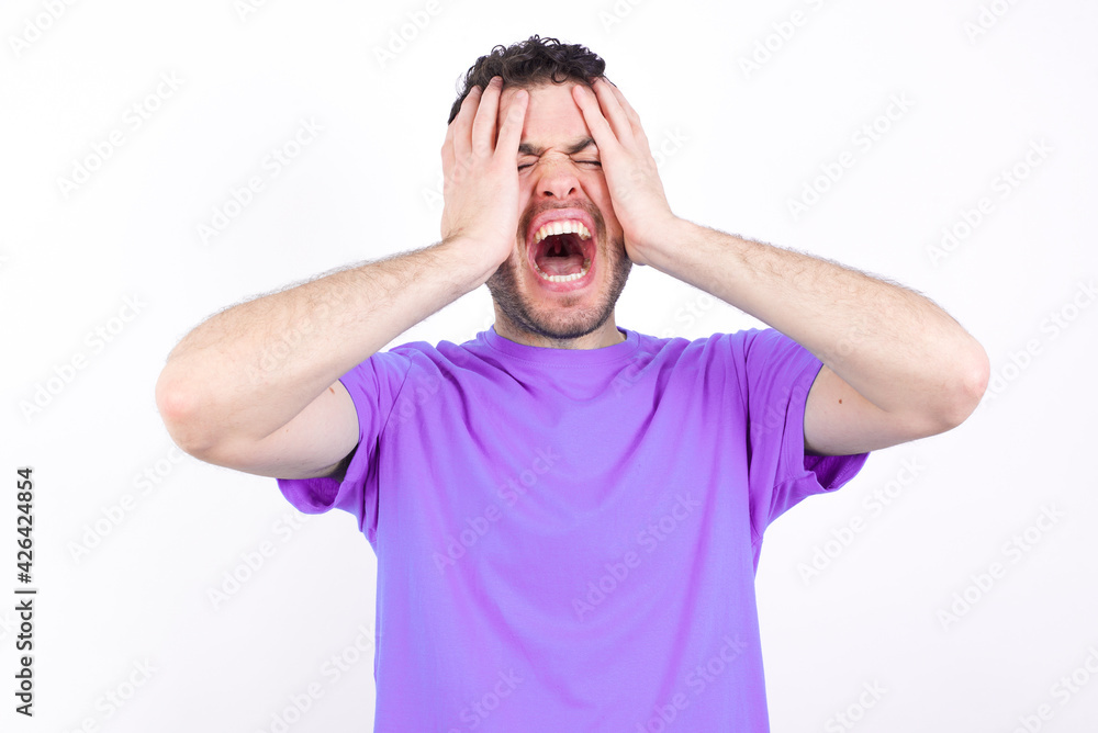 Shocked panic young handsome caucasian man wearing purple t-shirt against white background holding hands on head and screaming in despair and frustration. Shocked panic young handsome caucasian man wearing purple t-shirt against white background holding hands on head and screaming in despair and frustration.