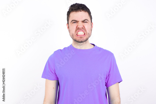 Mad crazy young handsome caucasian man wearing purple t-shirt against white background clenches teeth angrily, being annoyed with coming noise. Negative feeling concept.