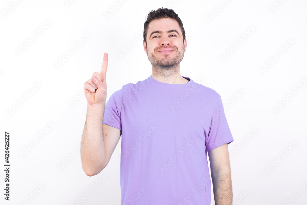 Naklejka premium young handsome caucasian man wearing purple t-shirt against white background showing and pointing up with fingers number one while smiling confident and happy.