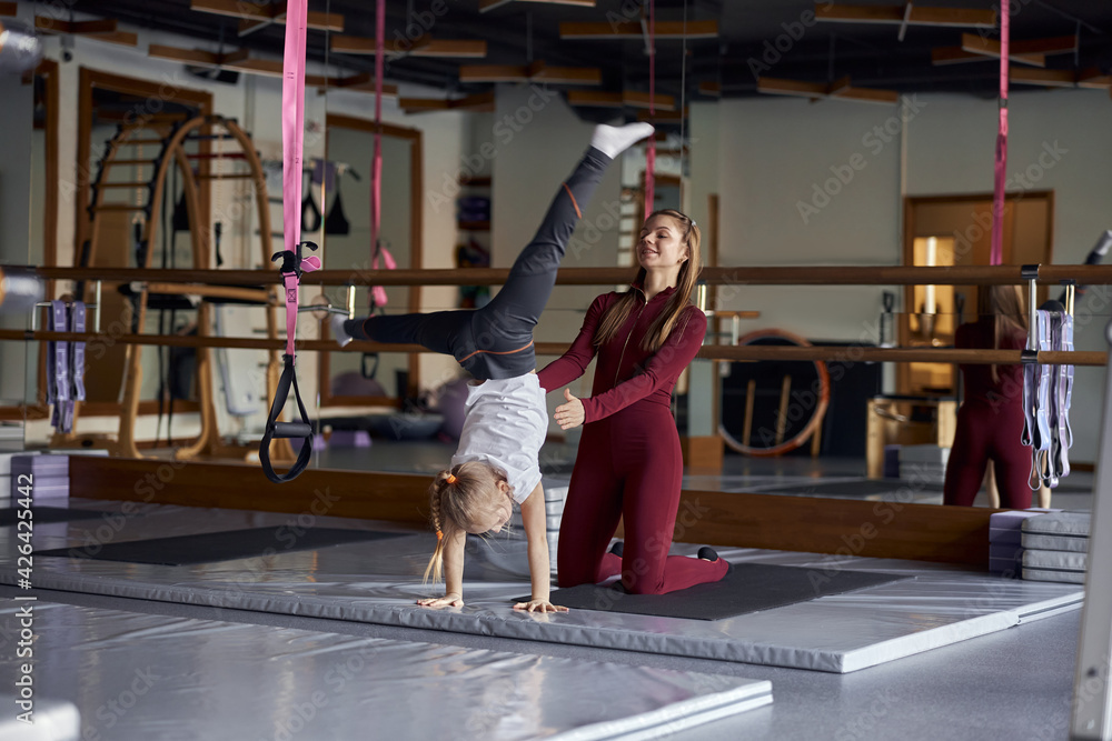 Young gymtastics teacher is helping a girl to do exercises indoors