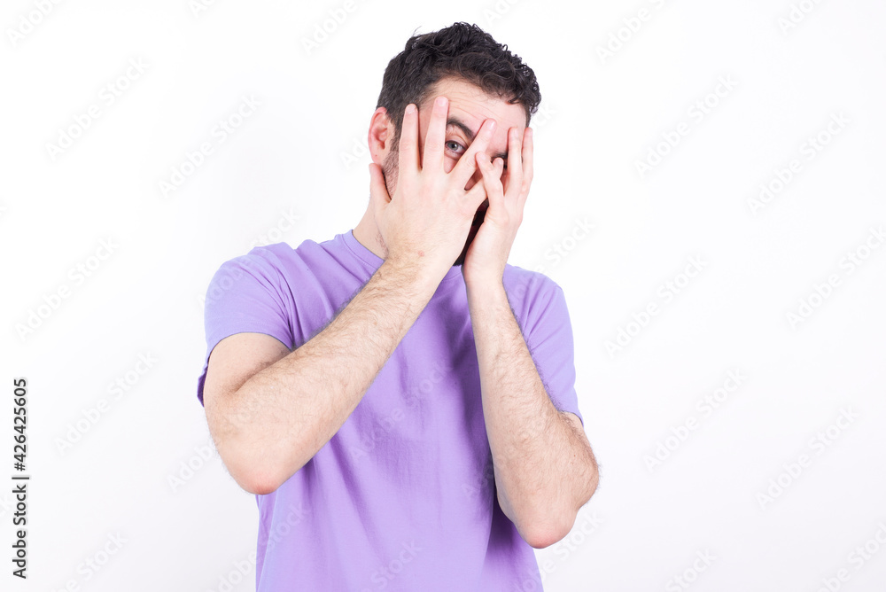 young handsome caucasian man wearing purple t-shirt against white background covering face with hands and peering out with one eye between fingers. Scared from something or someone.