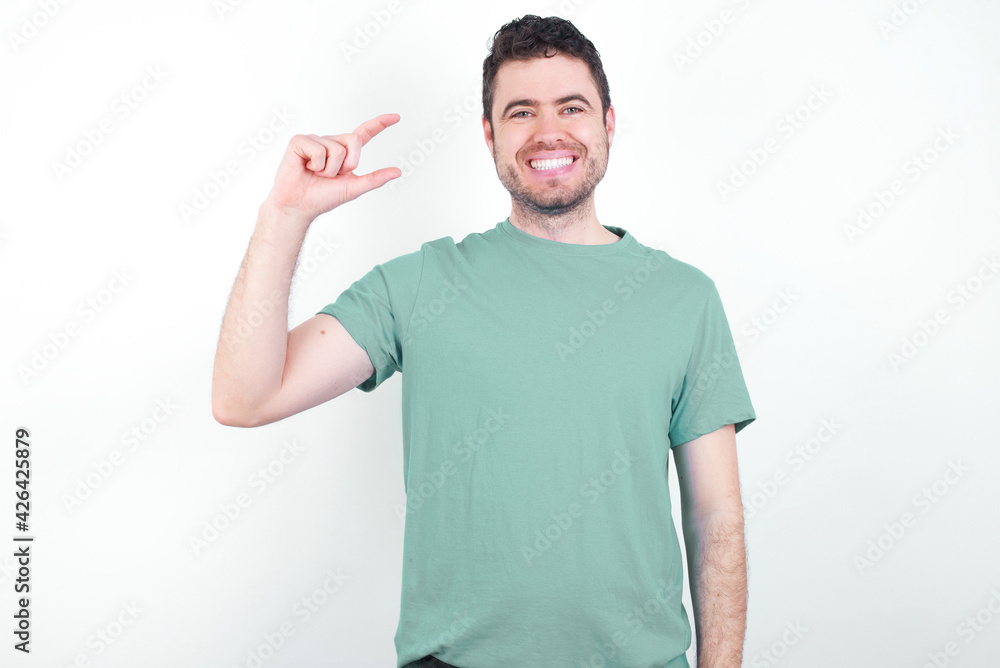 young handsome caucasian man wearing green t-shirt against white background smiling and gesturing with hand small size, measure symbol. young handsome caucasian man wearing green t-shirt against white background smiling and gesturing with hand small size, measure symbol.