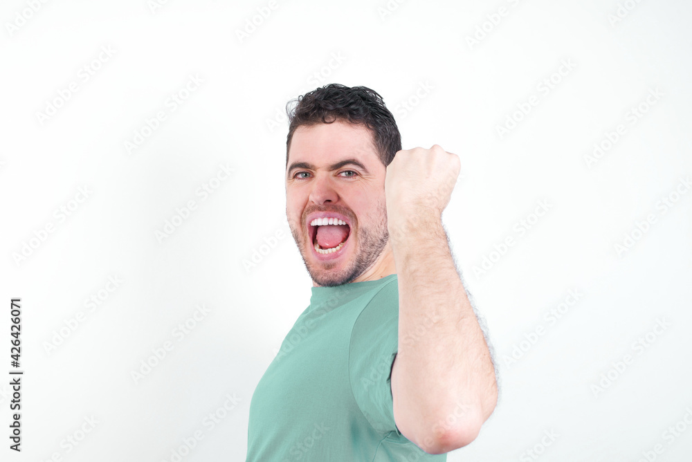 Overjoyed young handsome caucasian man wearing green t-shirt against white background glad to receive good news, clenching fist and making winning gesture. Overjoyed young handsome caucasian man wearing green t-shirt against white background glad to receive good news, clenching fist and making winning gesture.