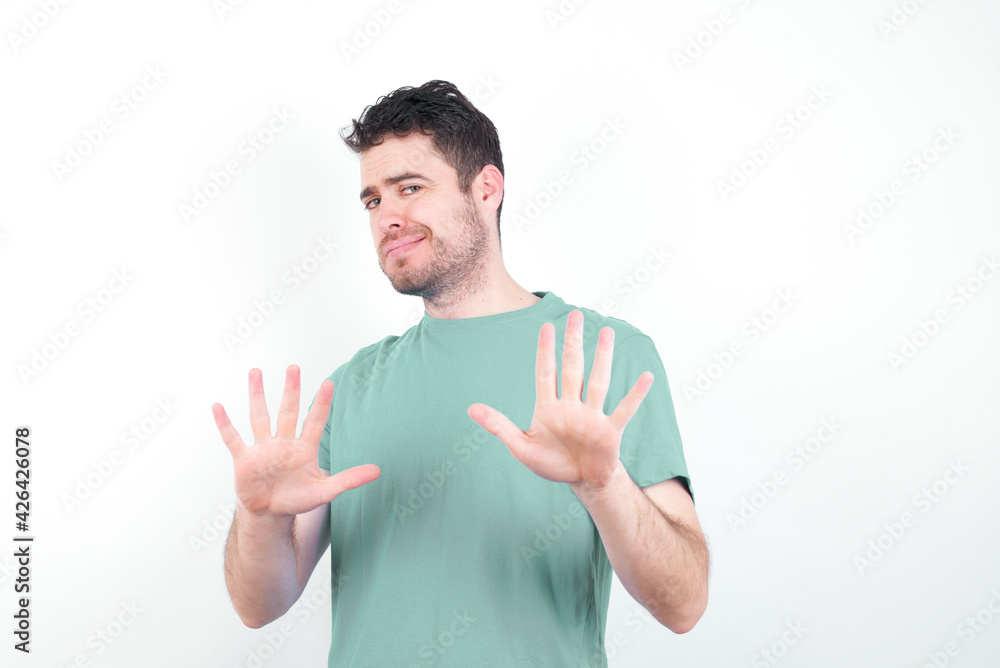 Afraid young handsome caucasian man wearing green t-shirt against white background, makes terrified expression and stop gesture with both hands saying: Stay there. Panic concept. Afraid young handsome caucasian man wearing green t-shirt against white background, makes terrified expression and stop gesture with both hands saying: Stay there. Panic concept.