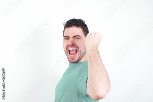 Overjoyed young handsome caucasian man wearing green t-shirt against white background glad to receive good news, clenching fist and making winning gesture.