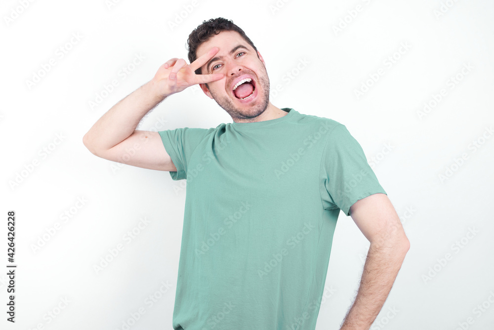 young handsome caucasian man wearing green t-shirt against white background making v-sign near eyes. Leisure, coquettish, celebration, and flirt concept. young handsome caucasian man wearing green t-shirt against white background making v-sign near eyes. Leisure, coquettish, celebration, and flirt concept.