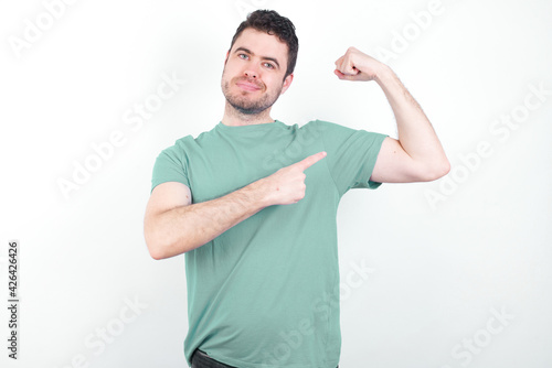 Smiling young handsome caucasian man wearing green t-shirt against white background raises hand to show muscles, feels confident in victory, strong and independent.