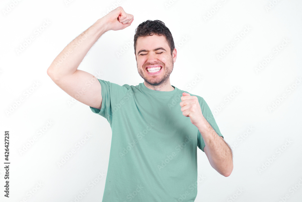 Attractive young handsome caucasian man wearing green t-shirt against white background celebrating a victory punching the air with his fists and a beaming toothy smile. Attractive young handsome caucasian man wearing green t-shirt against white background celebrating a victory punching the air with his fists and a beaming toothy smile.
