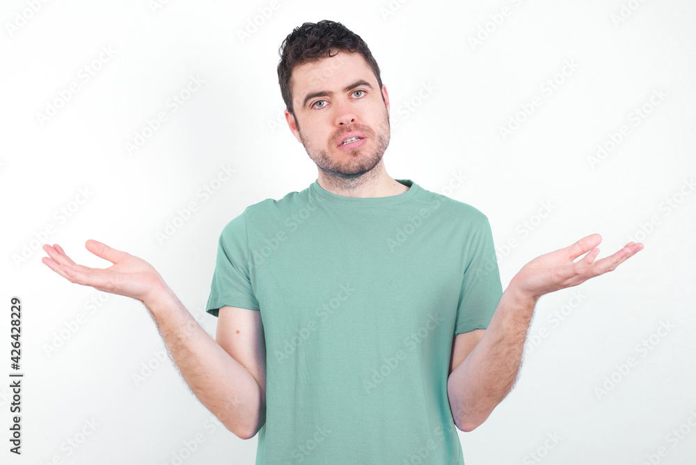 young handsome caucasian man wearing green t-shirt against white background looks uncertain shrugs shoulders. young handsome caucasian man wearing green t-shirt against white background looks uncertain shrugs shoulders.