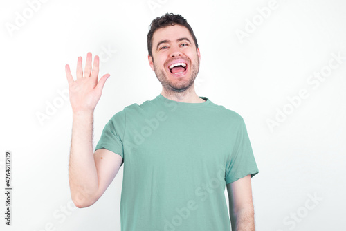 young handsome caucasian man wearing green t-shirt against white background waiving saying hello or goodbye happy and smiling, friendly welcome gesture.
