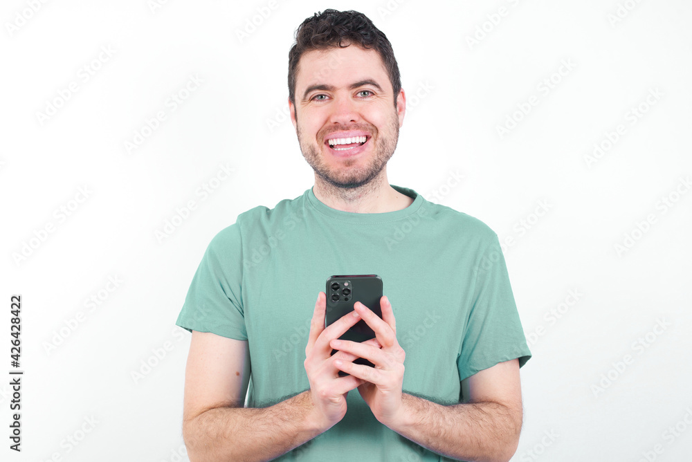 Smiling young handsome caucasian man wearing green t-shirt against white background friendly and happily holding mobile phone taking selfie in mirror. Smiling young handsome caucasian man wearing green t-shirt against white background friendly and happily holding mobile phone taking selfie in mirror.