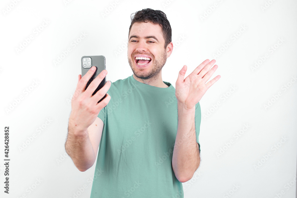 Portrait of happy friendly young handsome caucasian man wearing green t-shirt against white background taking selfie and waving hand, communicating on video call, online chatting. Portrait of happy friendly young handsome caucasian man wearing green t-shirt against white background taking selfie and waving hand, communicating on video call, online chatting.