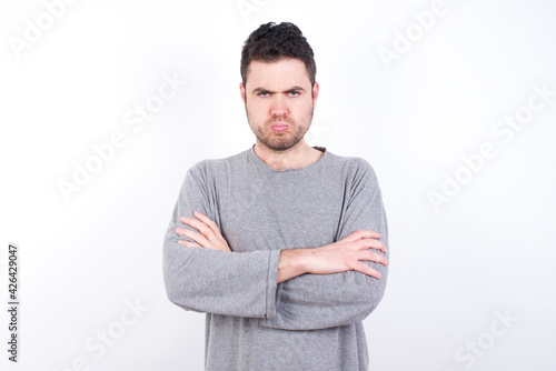 Gloomy dissatisfied young handsome caucasian bearded man wearing pyjama over white wall looks with miserable expression at camera from under forehead, makes unhappy grimace