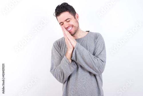 Overjoyed young handsome caucasian bearded man wearing pyjama over white wall laughs joyfully and keeps palms pressed together hears something funny