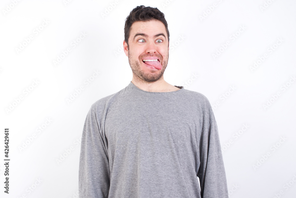 Obraz premium Funny young handsome caucasian bearded man wearing pyjama over white wall makes grimace and crosses eyes plays fool has fun alone sticks out tongue.