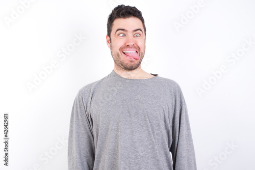 Funny young handsome caucasian bearded man wearing pyjama over white wall makes grimace and crosses eyes plays fool has fun alone sticks out tongue.