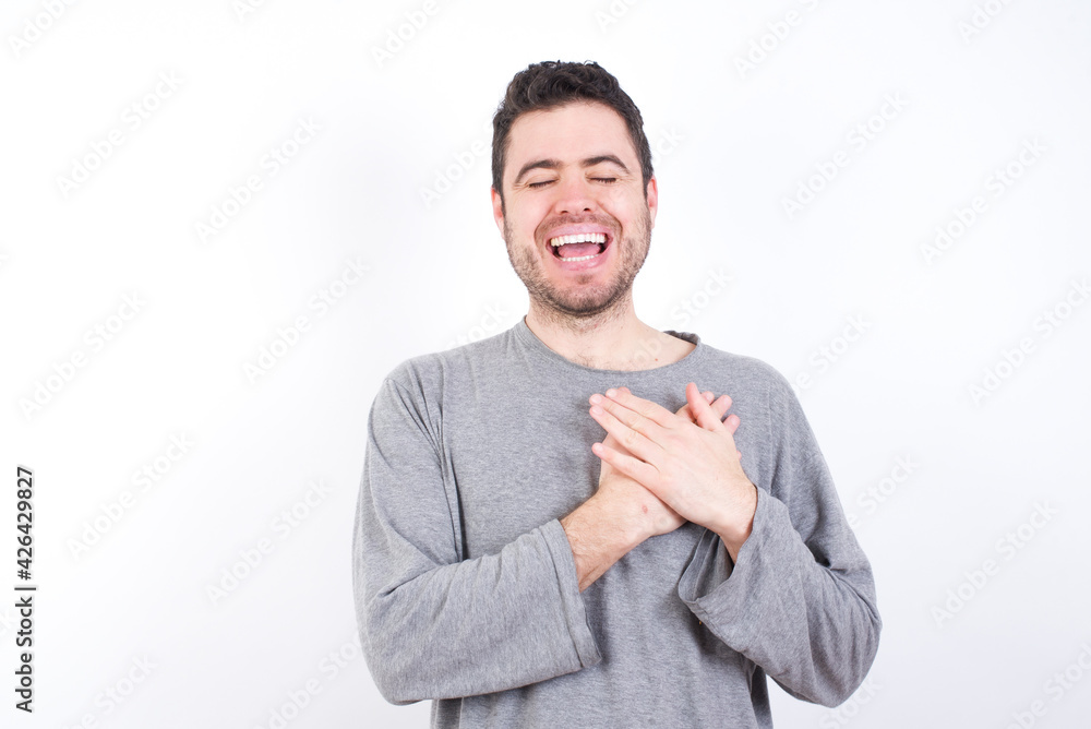 young handsome caucasian bearded man wearing pyjama over white wall expresses happines, laughs pleasantly, keeps hands on heart young handsome caucasian bearded man wearing pyjama over white wall expresses happines, laughs pleasantly, keeps hands on heart