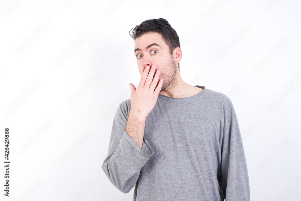 young handsome caucasian bearded man wearing pyjama over white wall covers mouth and looks with wonder at camera, cannot believe unexpected rumors. young handsome caucasian bearded man wearing pyjama over white wall covers mouth and looks with wonder at camera, cannot believe unexpected rumors.