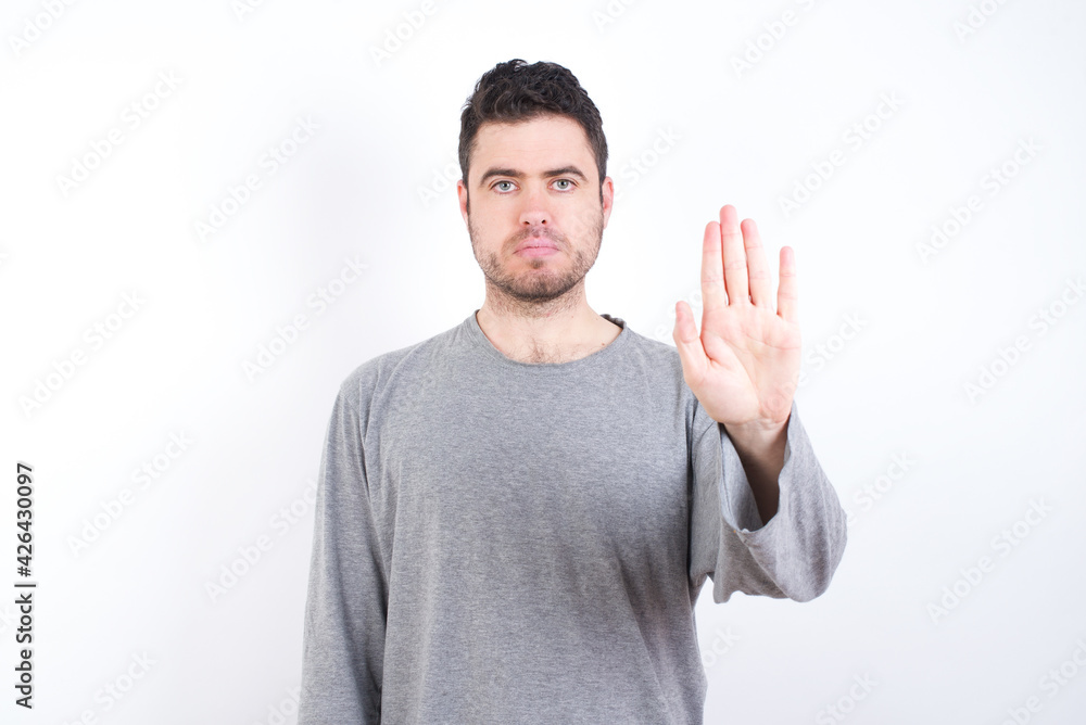 young handsome caucasian bearded man wearing pyjama over white wall shows stop sign prohibition symbol keeps palm forward to camera with strict expression young handsome caucasian bearded man wearing pyjama over white wall shows stop sign prohibition symbol keeps palm forward to camera with strict expression