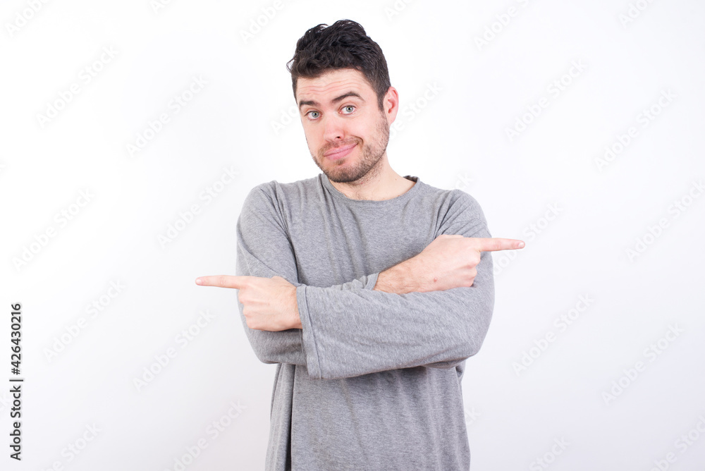 young handsome caucasian bearded man wearing pyjama over white wall crosses arms and points at different sides hesitates between two items or variants. Needs help with decision