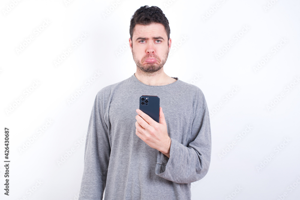 Upset dissatisfied young handsome caucasian bearded man wearing pyjama over white wall uses mobile software application and surrfs information in internet, holds modern mobile hand Upset dissatisfied young handsome caucasian bearded man wearing pyjama over white wall uses mobile software application and surrfs information in internet, holds modern mobile hand