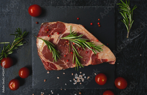 Raw piece of ribeye beef meat on a slate board on a wooden table with tomato, basil and himalayan salt. Fresh food. Everything for a barbecue.