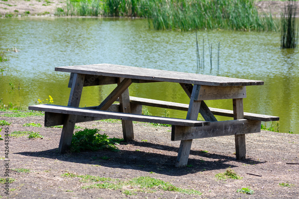 Empty wooden picnic table with attached benches set up next to the shallow pond covered covered by reeds in the public park. Empty wooden picnic table with attached benches set up next to the shallow pond covered covered by reeds in the public park.