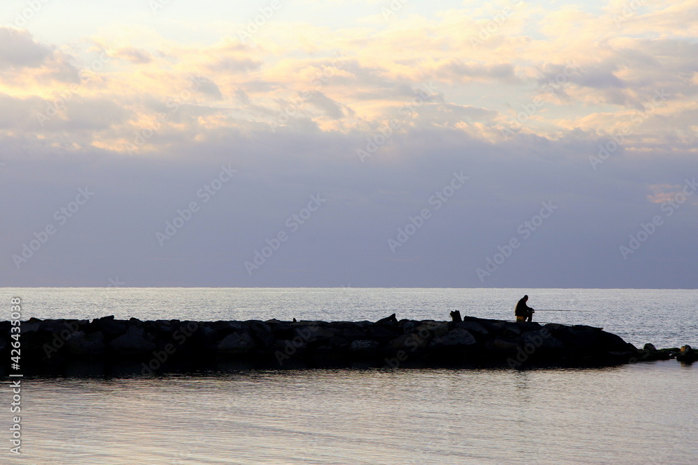 Silhouette of a fisherman sitting with his fishing rod on the rocks in the sunset light over the sea