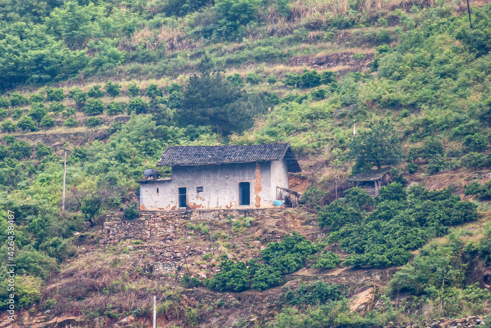 Three Gorges Dam, China - May 6, 2010: Closeup of small white farm building with dark gray roof set on green hill slope with terraced agriculture. Patches of brown dirt.