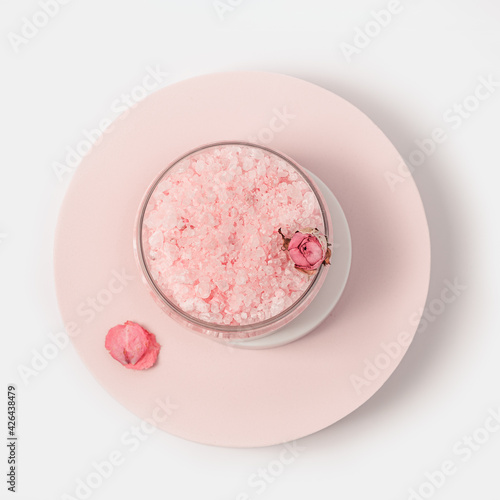 Rose-scented beauty sea salt in jar with cap and rose flowers on round podium. Cosmetic product top view, pink pastel colored