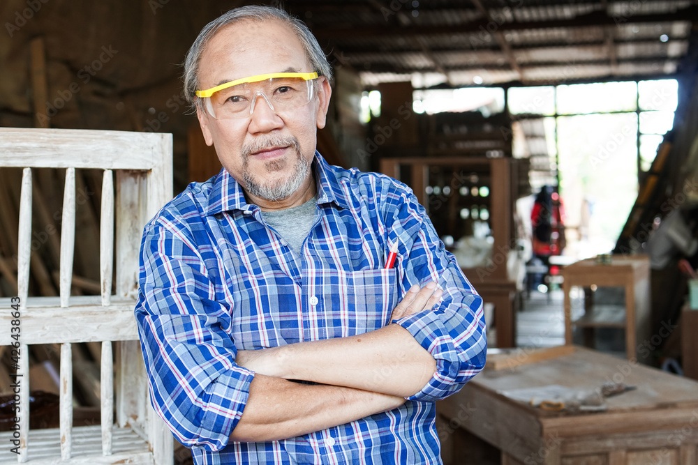 Portrait of Asian senior male carpenter in arms crossed pose in the workshop with wood boards background. copy space Portrait of Asian senior male carpenter in arms crossed pose in the workshop with wood boards background. copy space