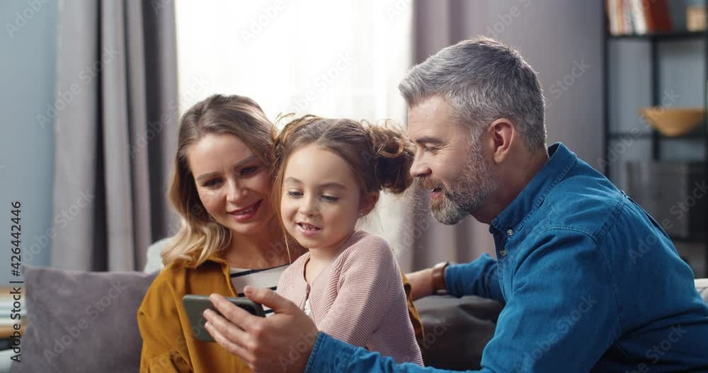 Happy mom and dad sit at home on couch with their little daughter and show her funny cartoon on smartphone. Happy mom and dad sit at home on couch with their little daughter and show her funny cartoon on smartphone.