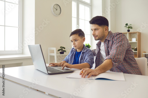 Caucasian father help son with online homework. Parent and schoolchild sitting at desk front of laptop in home living-room. Homeschooling during stay home and quarantine in virus outbreak