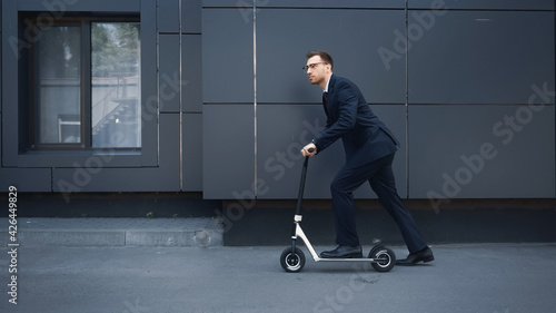 full length of businessman in glasses and suit riding e-scooter near building.