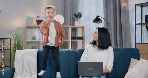 Beautiful smiling happy mother with black hair admiring of her cute 2-aged son which jumping near her on couch and then hugging him photo