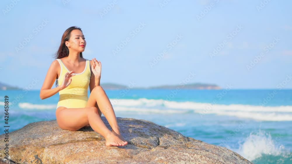 Beautiful Asian woman in yellow monokini sitting on a big rock on the seafront at Caribbean island and touching her hair, slow-motion handheld
