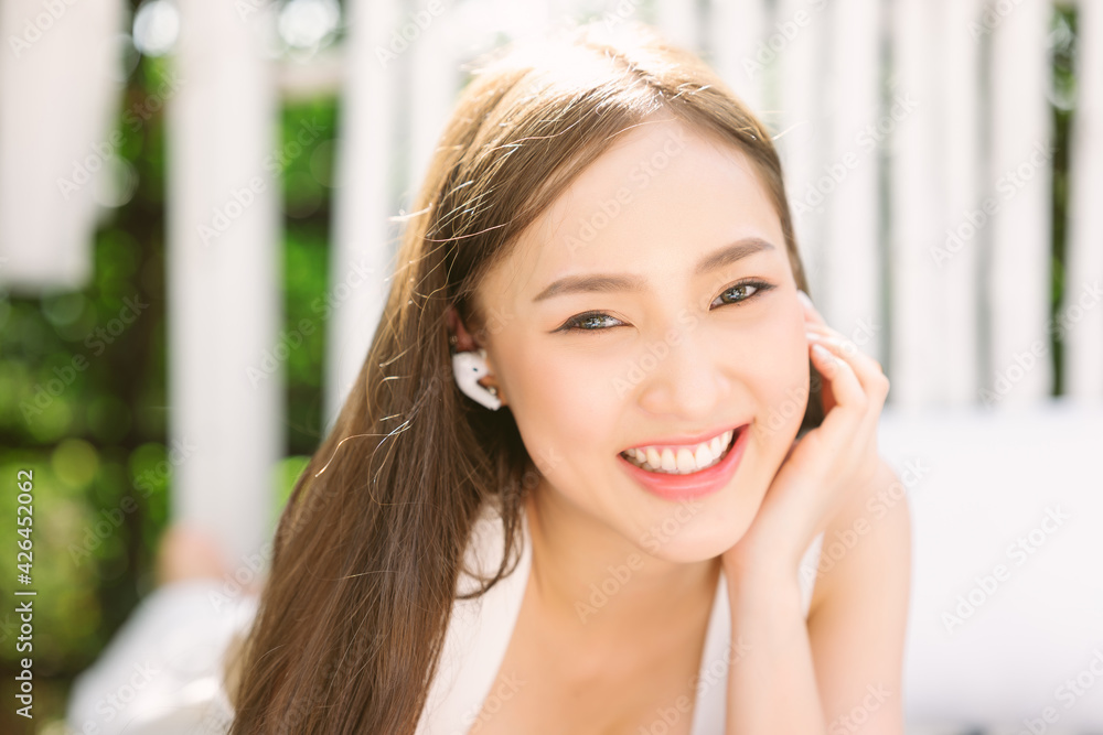 Close-up Portrait of smiling young Asian woman touching wireless earbud, listening music in headphone and looking at camera while resting on bench in park. Weekend Relaxation Leisure Concept.