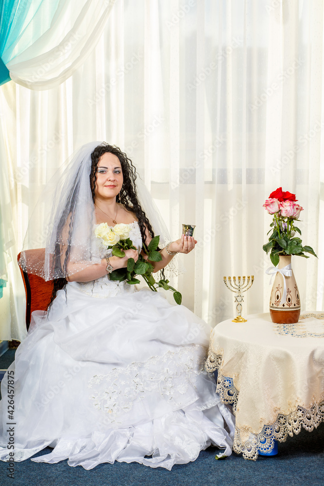 A happy Jewish bride sits in the hall before the chuppah ceremony at a table with flowers, a glass of wine and a bouquet of white roses. A happy Jewish bride sits in the hall before the chuppah ceremony at a table with flowers, a glass of wine and a bouquet of white roses.
