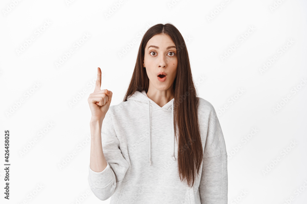 Impressed girl telling about promotional offer, pointing finger up, gasping and looking shocked at camera, say wow, recommending follow website, click on banner, white background Impressed girl telling about promotional offer, pointing finger up, gasping and looking shocked at camera, say wow, recommending follow website, click on banner, white background
