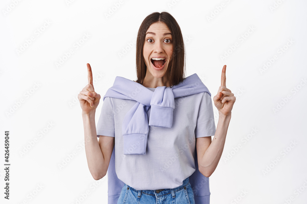 Amazing look there. Excited girl screams impressed, say wow and point up at promo deal, showing advertisement, standing over white background Amazing look there. Excited girl screams impressed, say wow and point up at promo deal, showing advertisement, standing over white background