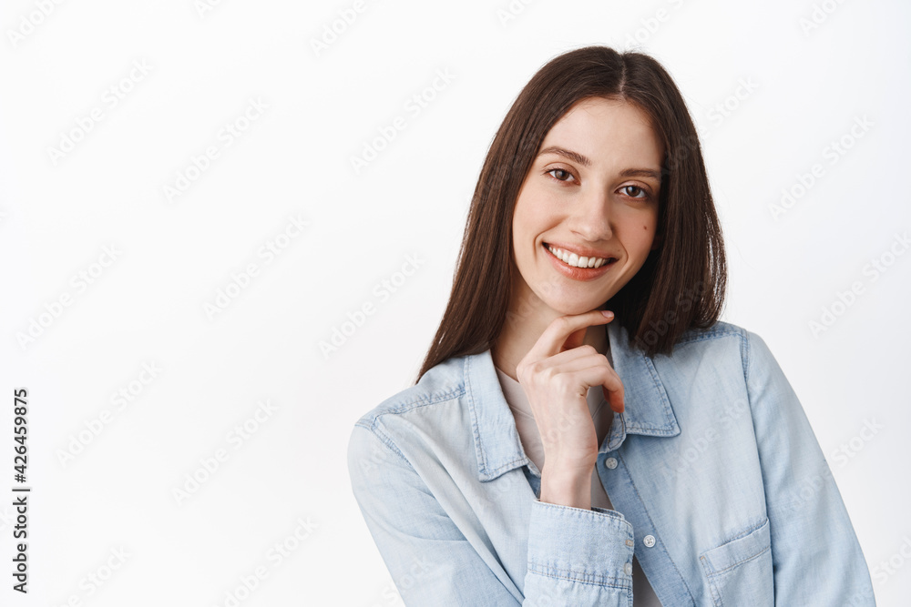 Close up of pensive smiling girl, touch chin, showing pure clean perfect skin, white smile teeth, standing against studio background Close up of pensive smiling girl, touch chin, showing pure clean perfect skin, white smile teeth, standing against studio background