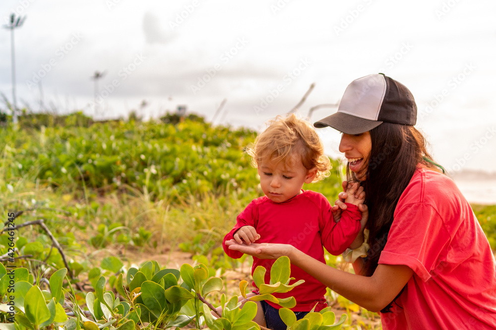 MÃE MOSTRANDO A VEGETAÇÃO DA PRAIA PARA O FILHO, COM O REFLEXO DO SOL ATRAS EM UMA BELA MANHÃ. MÃE MOSTRANDO A VEGETAÇÃO DA PRAIA PARA O FILHO, COM O REFLEXO DO SOL ATRAS EM UMA BELA MANHÃ.