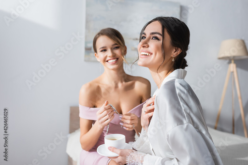 happy bride holding coffee cup and looking away near friend with necklace on blurred background.