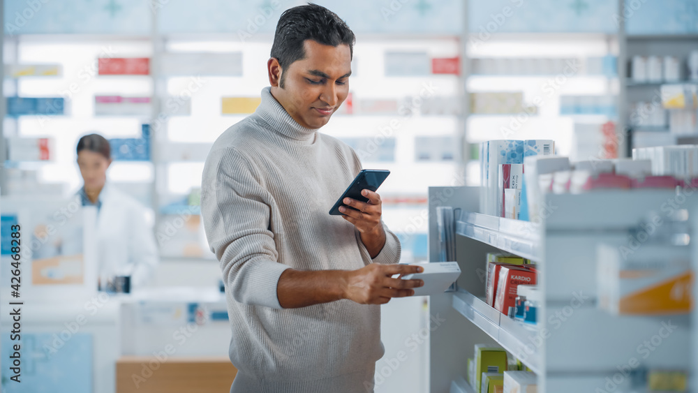 Pharmacy Drugstore: Portrait of a Handsome Young Indian Man Using Smartphone Device, Chooses to Purchase Best Medicine, Drugs, Vitamins. Shelves full of Sport Supplements, Health Care Products